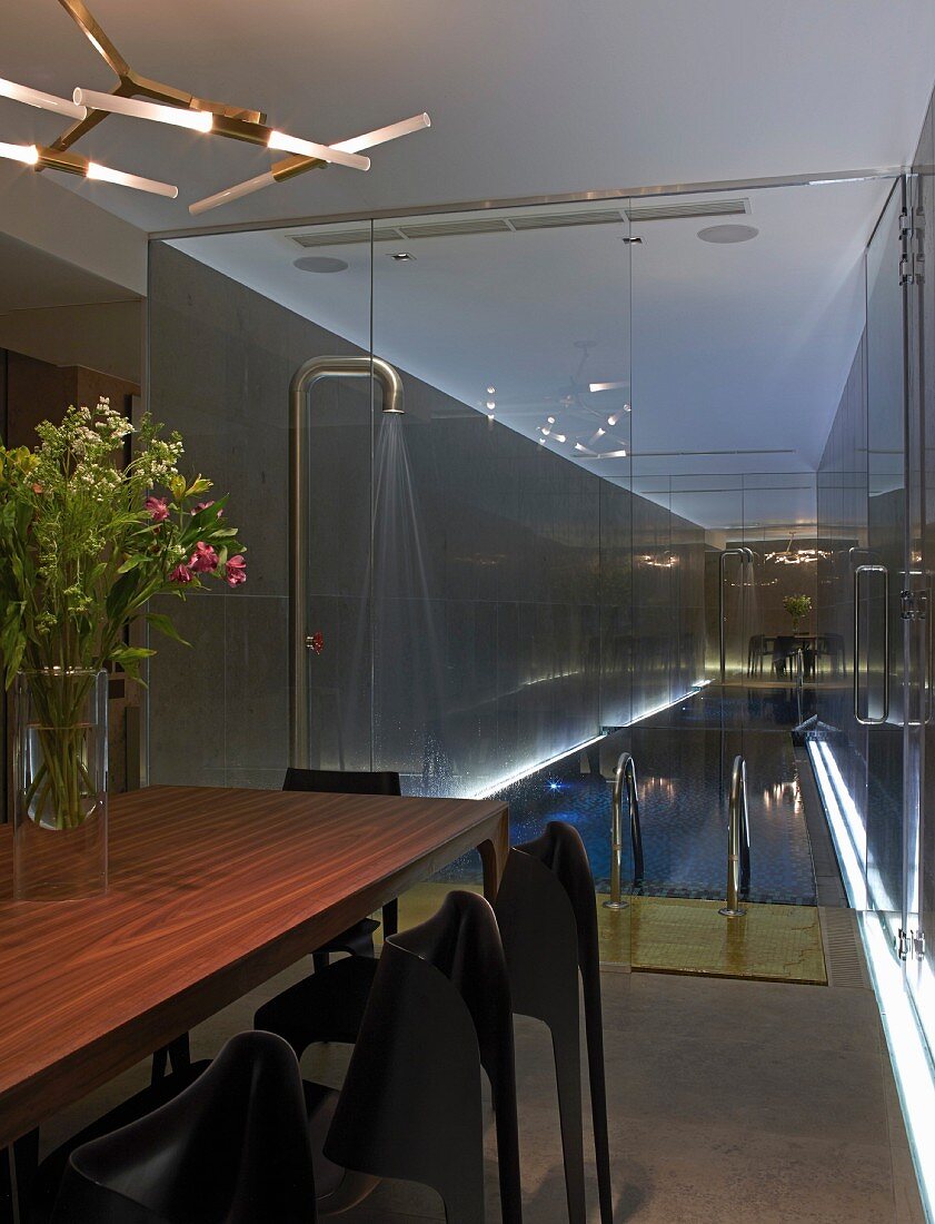 Edelholztisch mit Designerleuchte vor Glastrennwand und beleuchtetem Indoor-Pool mit Dusche