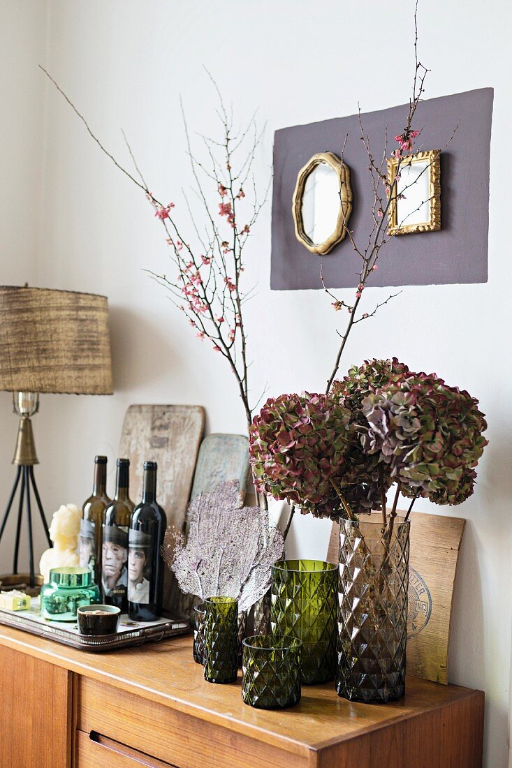 Glasvasen mit Blütenzweigen und Hortensien neben Weinflaschen auf Anrichte