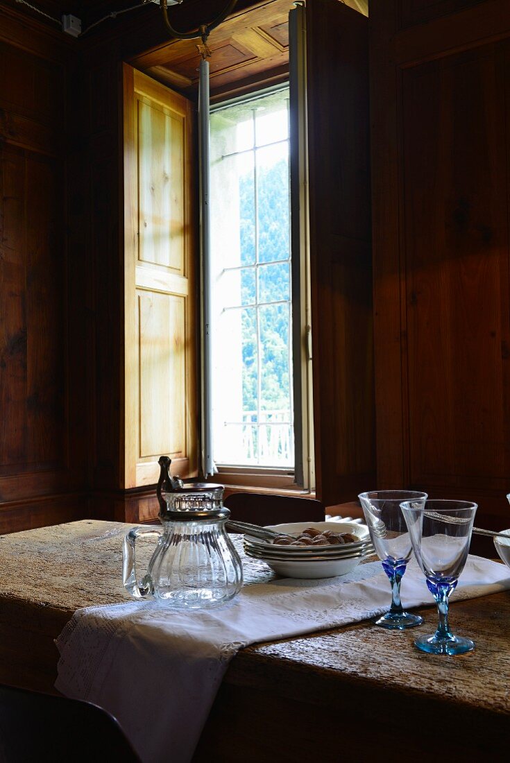 Glasses, carafe and plates on rustic dining table next to window in wood-clad dining room