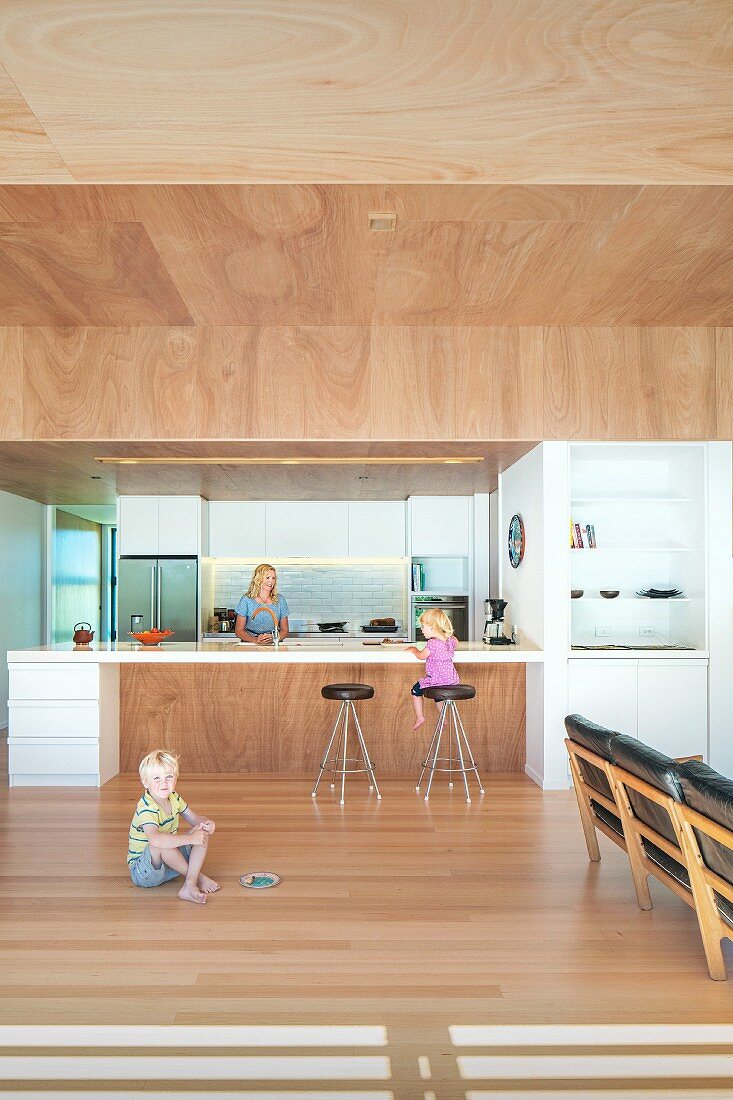 Children in open-plan interior and mother behind free-standing kitchen counter below suspended wooden ceiling, 