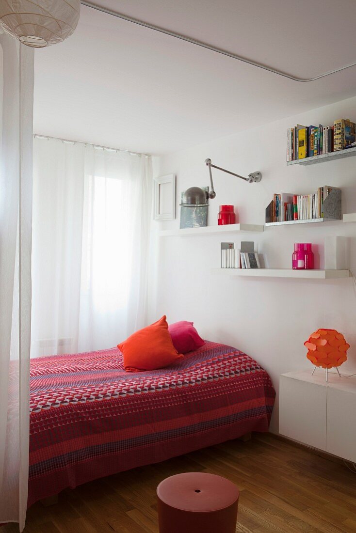 Bett mit Tagesdecke in Rottönen und folkloristischem Muster, weiße Wandboards und Hängeschrank
