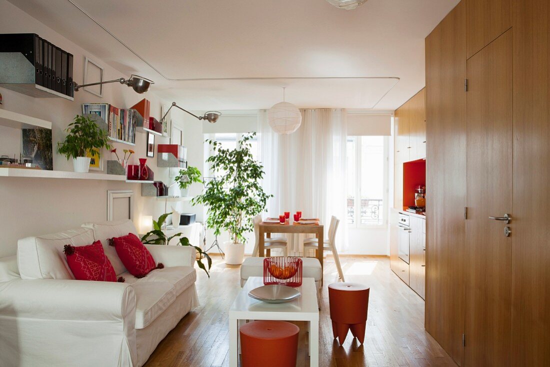 Einzimmerwohnung mit Einbauküche und orangefarbenen Hockern, weißem Hussensofa und Grünpflanze
