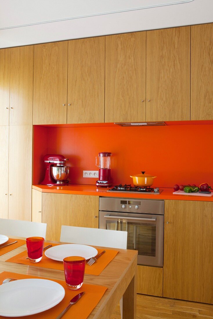 Gedeckter Esstisch vor Einbauküche mit Holzfronten und orangefarbenem Spritzschutz