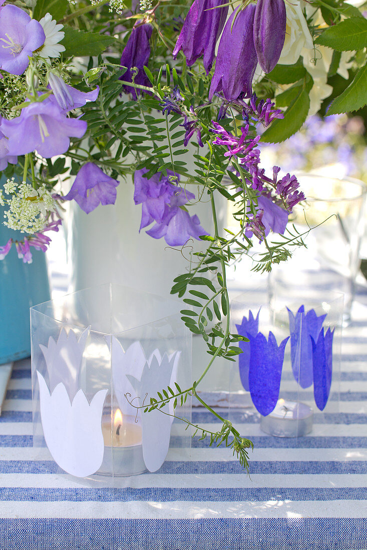 Candle lanterns decorated with paper bellflower silhouettes in front of vase of flowers