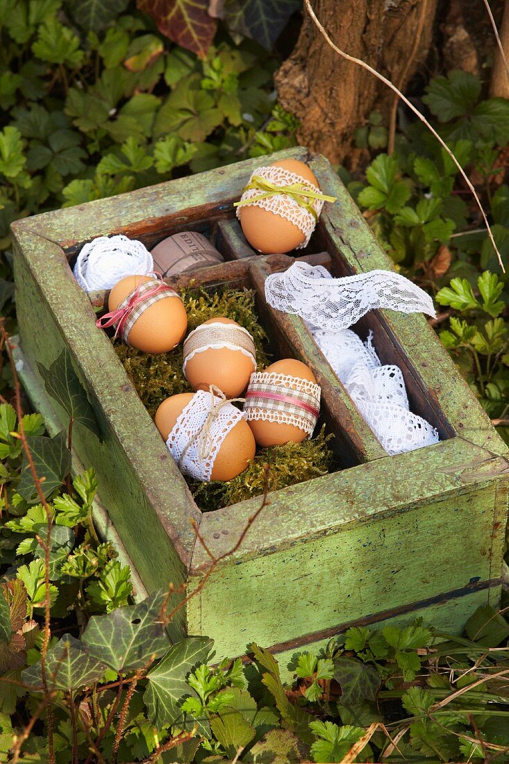 Undyed Easter eggs with lace trim and delicate satin ribbons on moss in vintage wooden crate