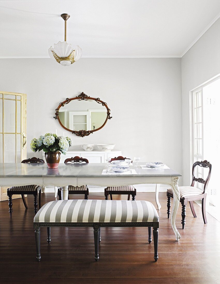 Upholstered bench and chairs in front of antique table in dining room with oval mirror on wall in background