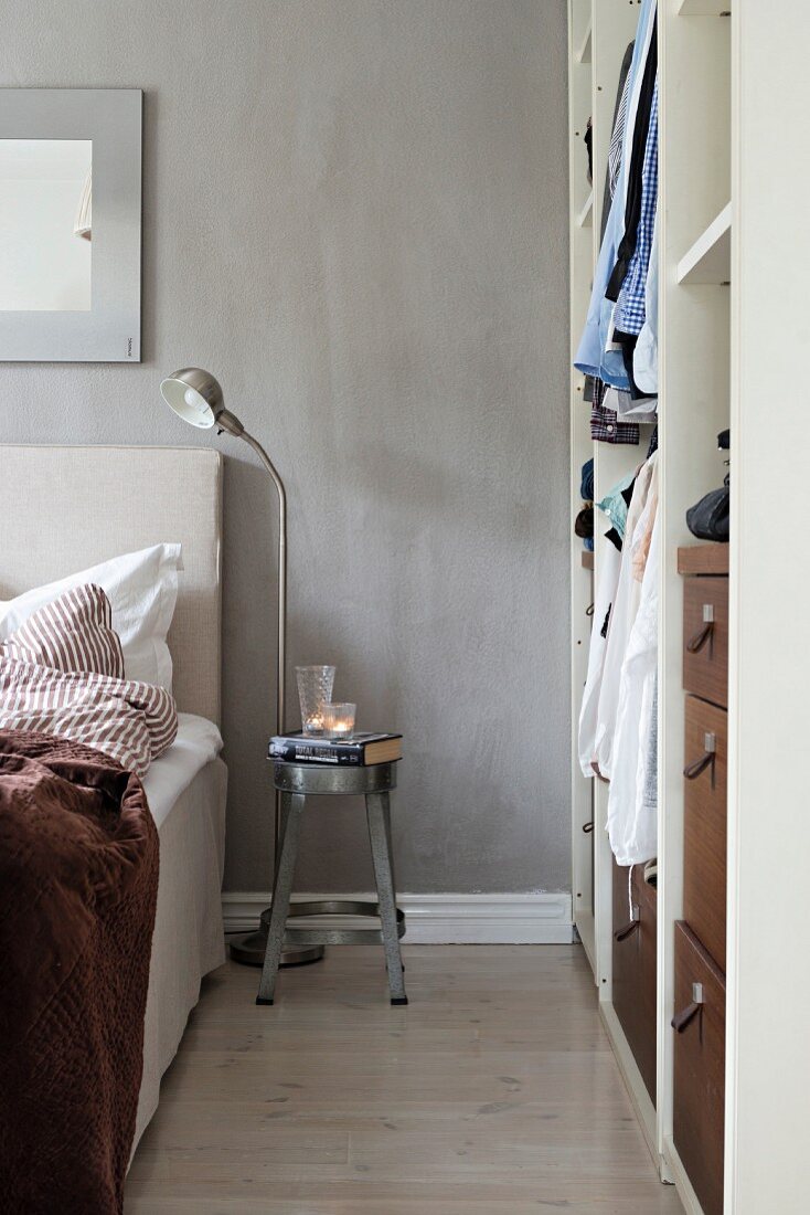 Metal stool used as bedside table against grey wall next to open-fronted cupboard