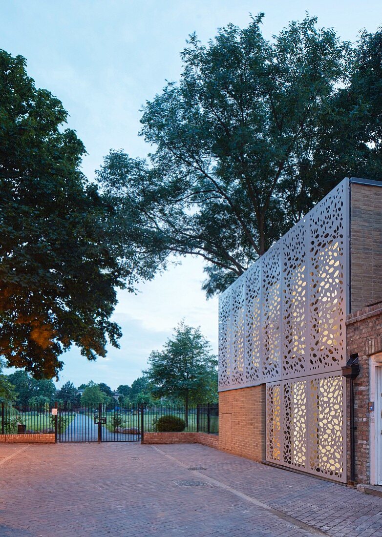 Building with modern perforated façade elements, paved courtyard and view into park-style gardens at twilight