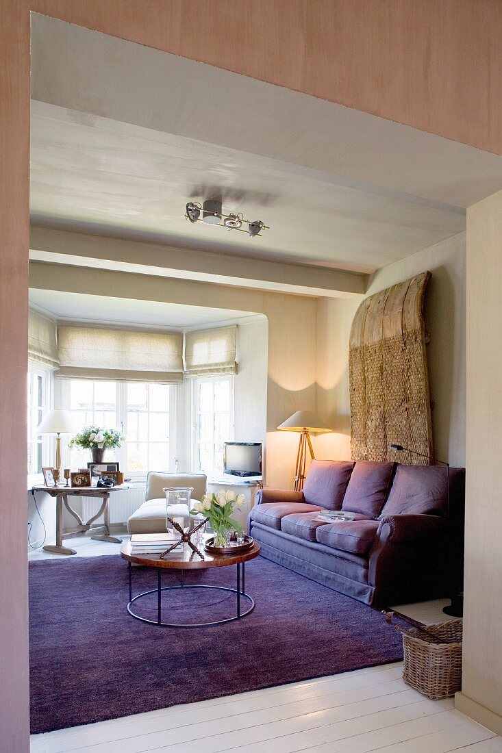Gemütlicher Loungebereich mit lilafarbener Couch und passendem Teppich vor hellem Erker