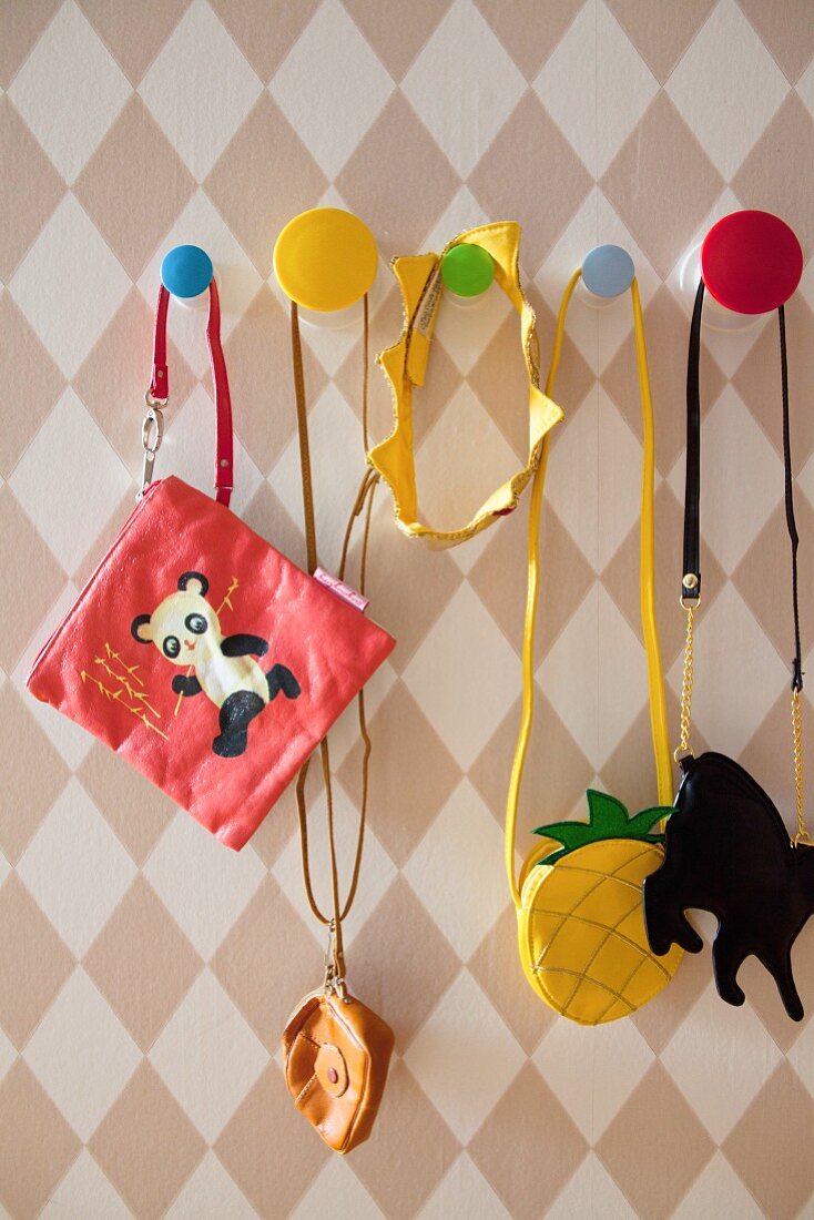 Various children's bags hung from colourful pegs on diamond-patterned wallpaper