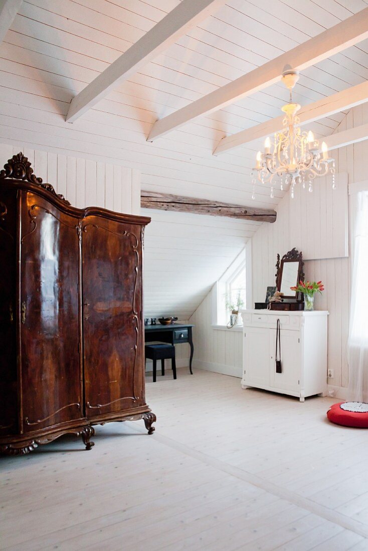 Antiker, kunsthandwerklicher Holzschrank in weißem Dachzimmer mit Kronleuchter und nostalgischem Flair