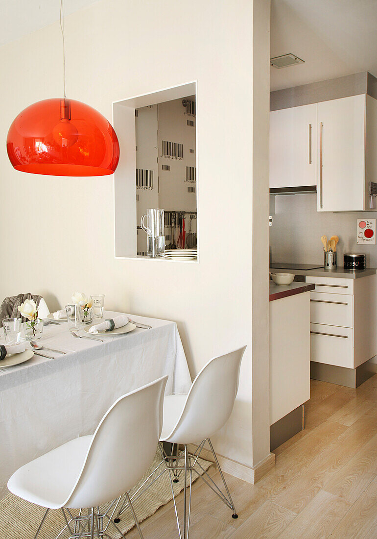 Dining table and white shell chairs below transparent red lampshade next to serving hatch