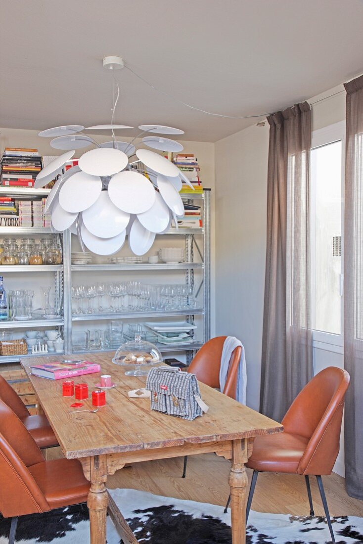 Rustic wooden table, leather chairs and white pendant lamp in front of crockery and books on metal shelves