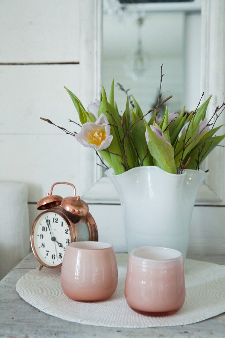 Rosafarbene Windlichter, ein Wecker und Tulpen in weißer Vase