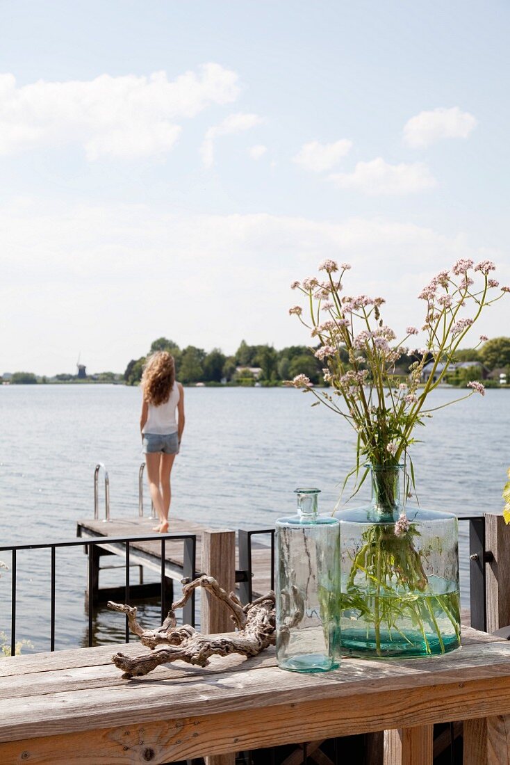 Glass bottle on wooden table next to lake with girl stood on jetty in background
