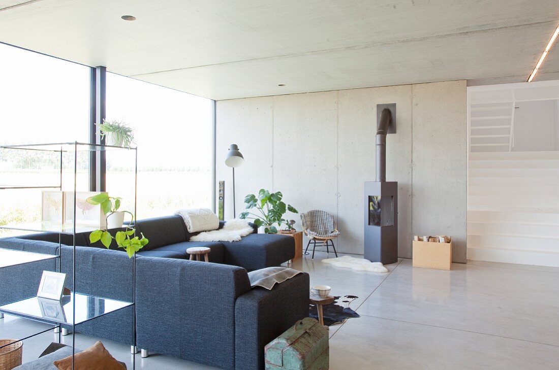 Open-sided glass shelves, grey couch and wood-burning stove in open-plan interior with glass wall and concrete wall