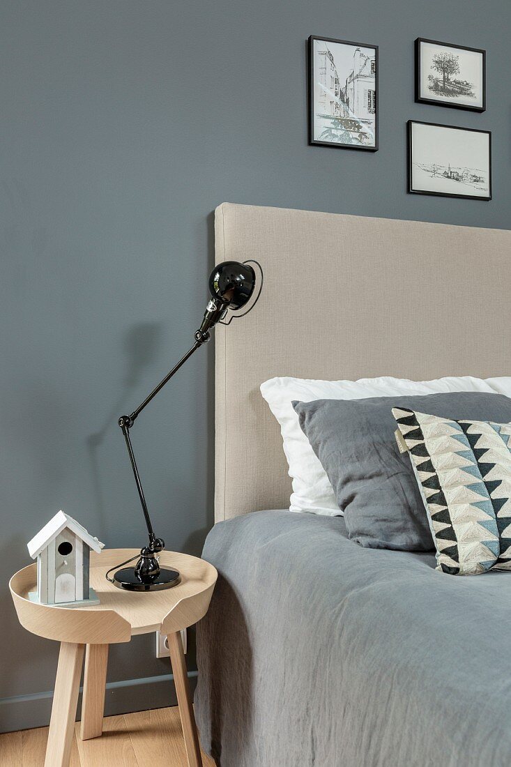 Bett mit beigefarbenem Kopfteil und grauer Decke an gleichfarbiger Wand, Nachttisch mit schwarzer Lampe