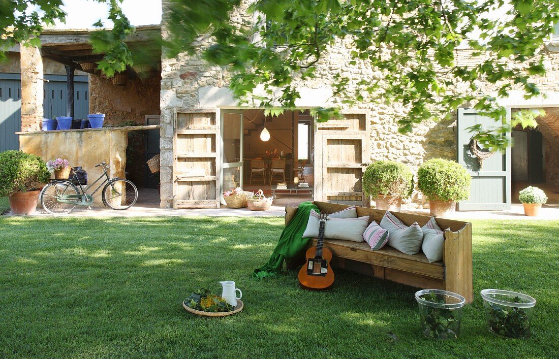 Holzbank mit Gitarre im Garten vor Natursteinhaus mit geöffneten Terrassentüren