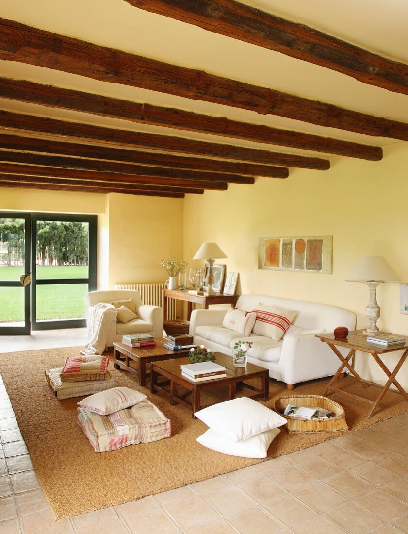 White sofa, floor cushions, side table and wood-beamed ceiling in comfortable lounge area of renovated country house