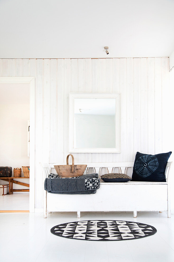 Cushions and wooden trug on white bench below mirror on white board wall