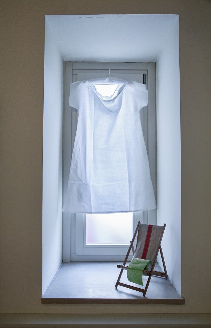 Simple white dress hung in window in place of curtain and miniature deckchair on windowsill