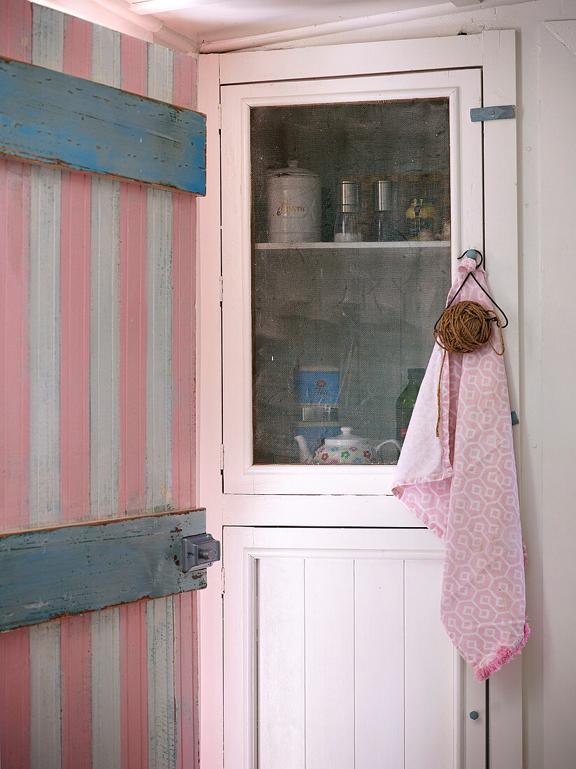 Blick durch rosa-weiss gestrichener Holztür auf Vitrinenschrank mit Küchenutensilien