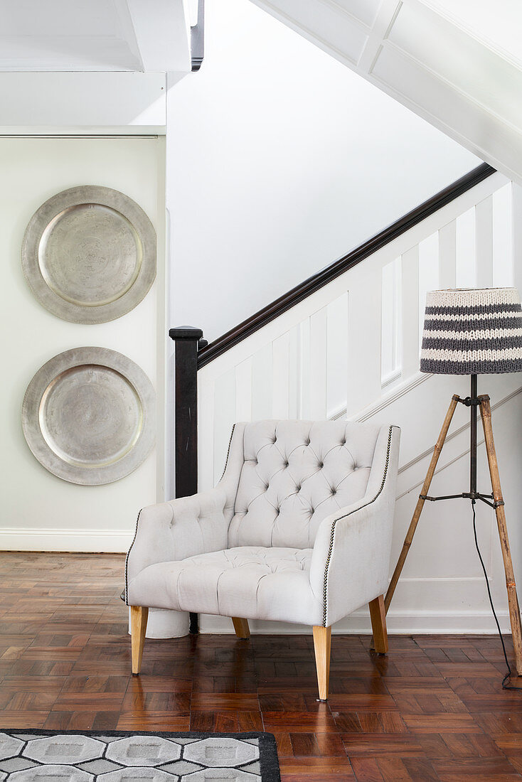 Armchair and standard lamp at foot of staircase with silver platters decorating wall in background