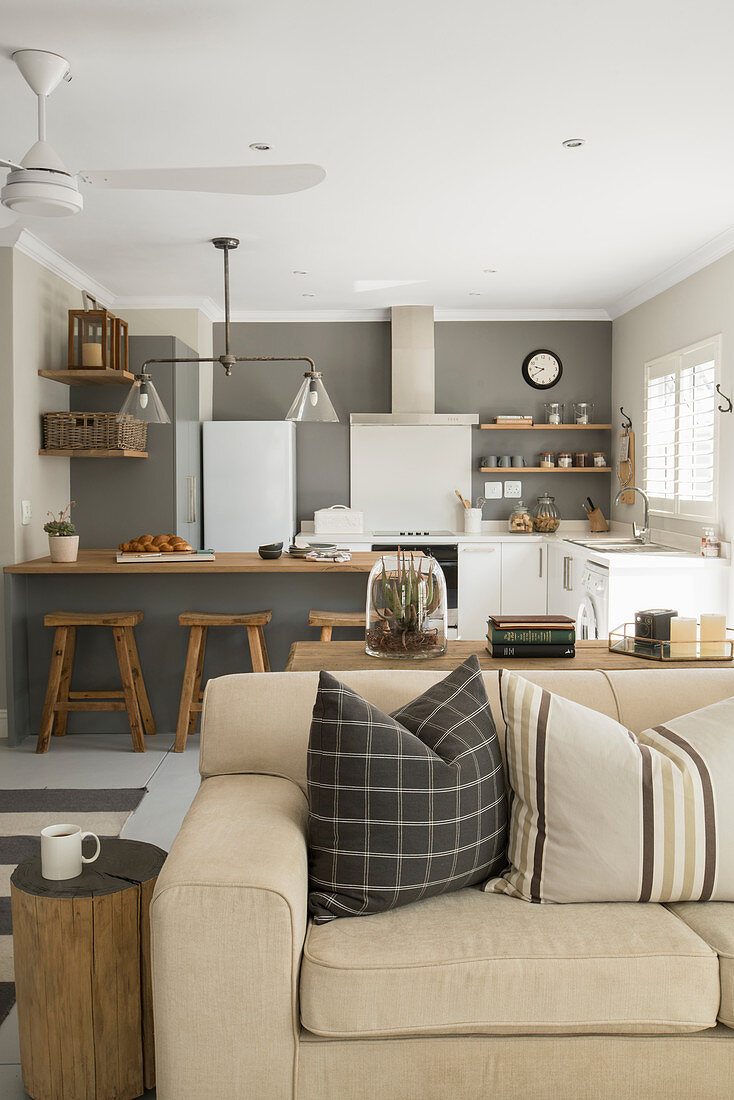 Helle Ausziehcouch mit Kissen und Baumstamm als Beistelltisch in offenem Wohnraum, im Hintergrund Küche mit Theke und gauer Wand