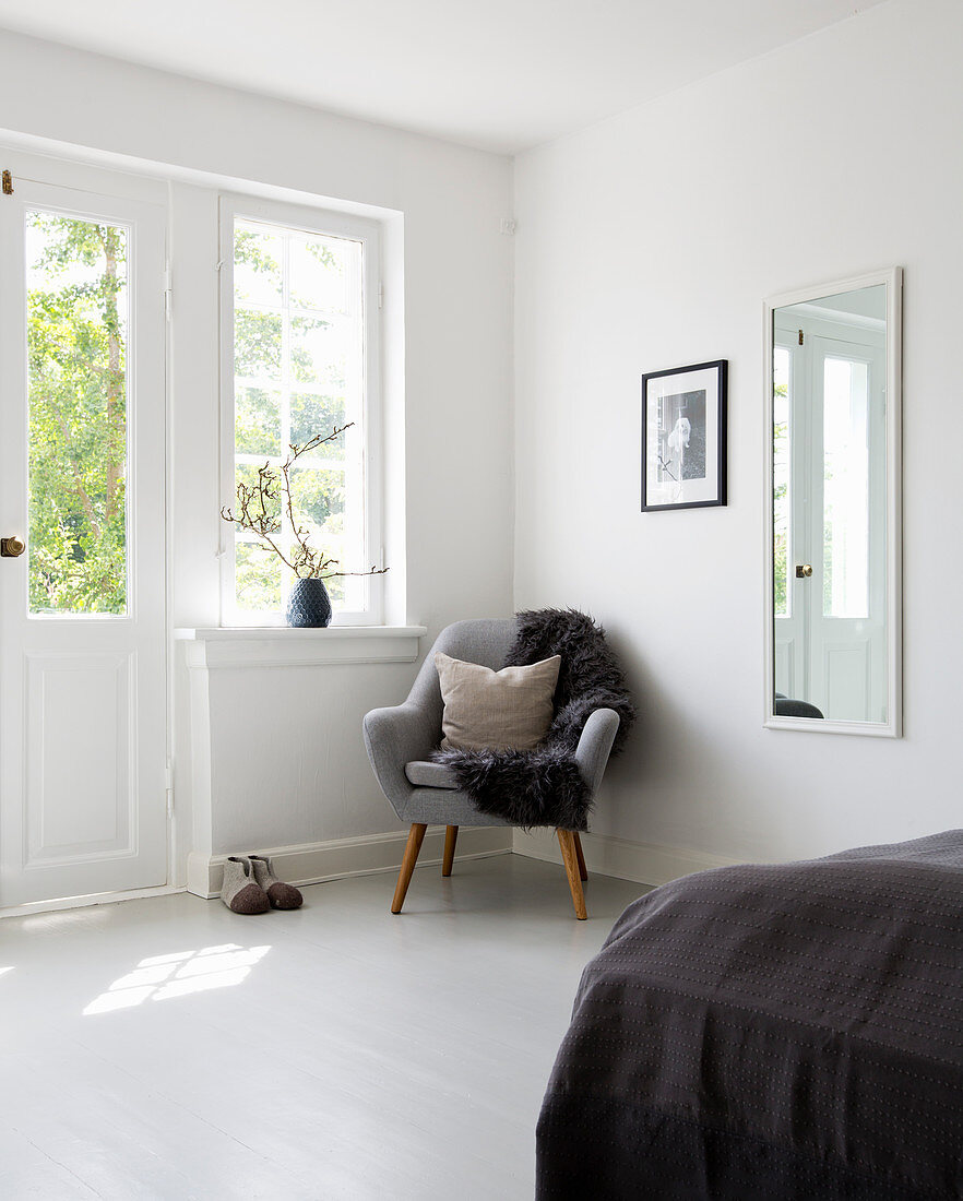Fur blanket and cushion on grey armchair below window in bedroom