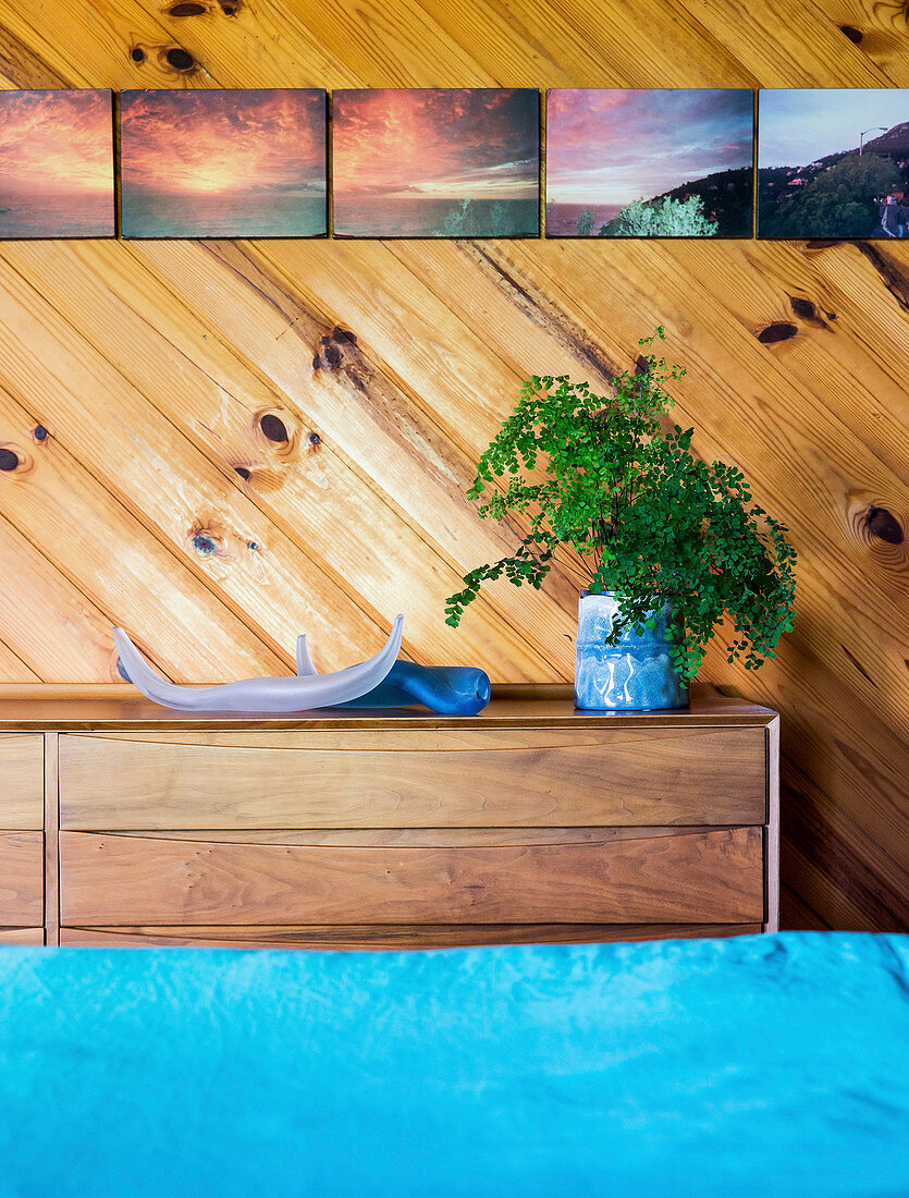Kommode vor Holzwand mit Bildersammlung im Schlafzimmer, im Vordergrund türkisfarbene Bettwäsche