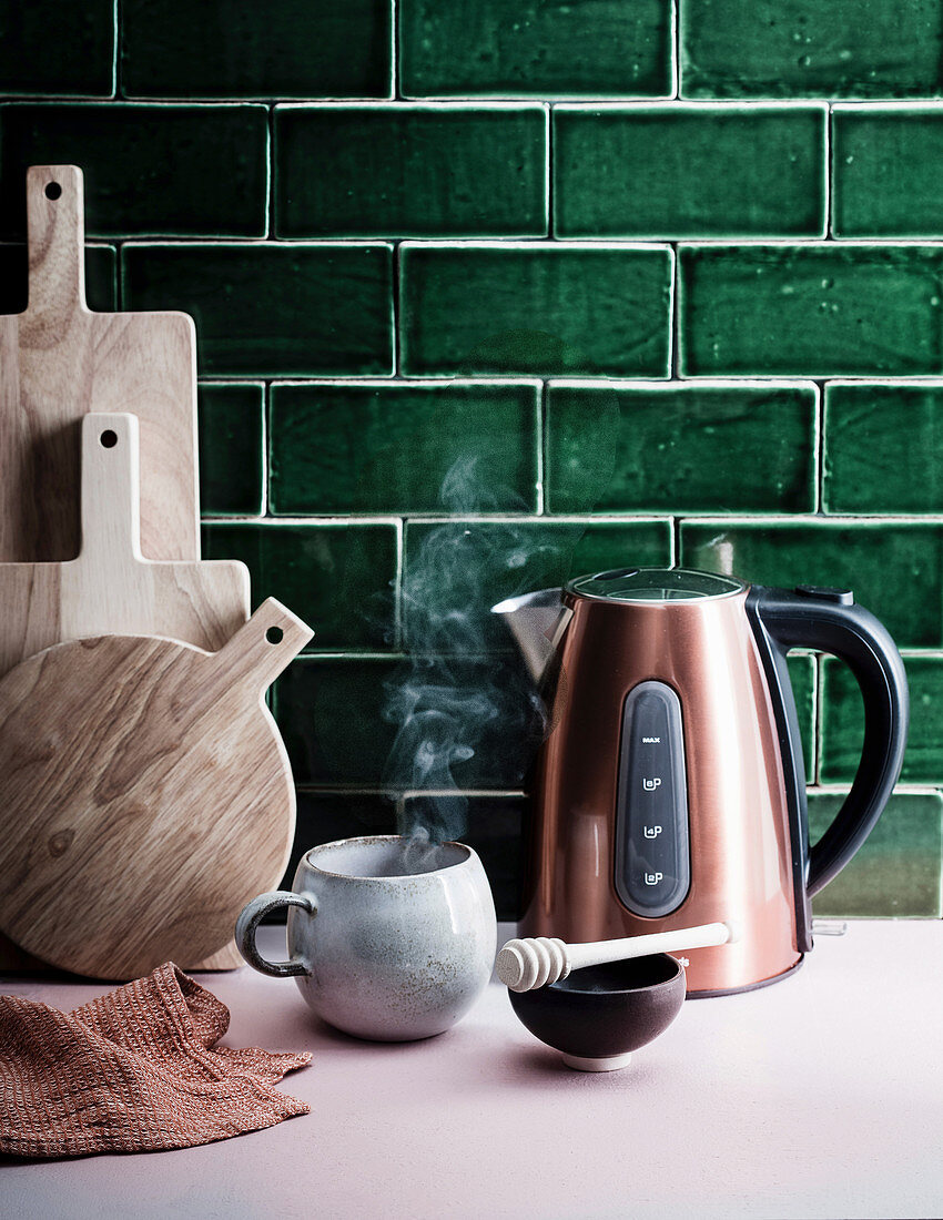 Kettle and a steaming cup in front of green tiles