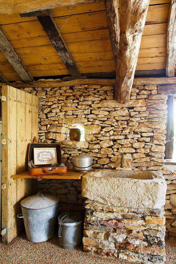 Suitcase and saucepan on wooden shelf next to stone basin in converted barn