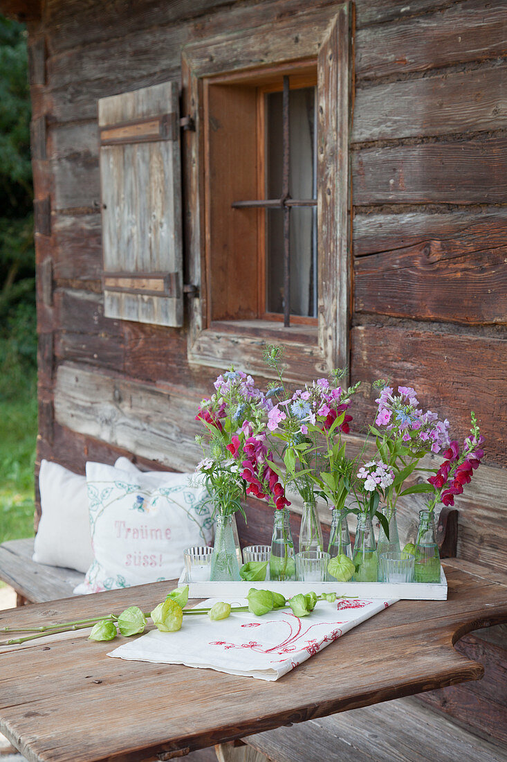 Phlox, love-in-a-mist and snapdragons in small bottles decorating table outside rustic wooden farmhouse