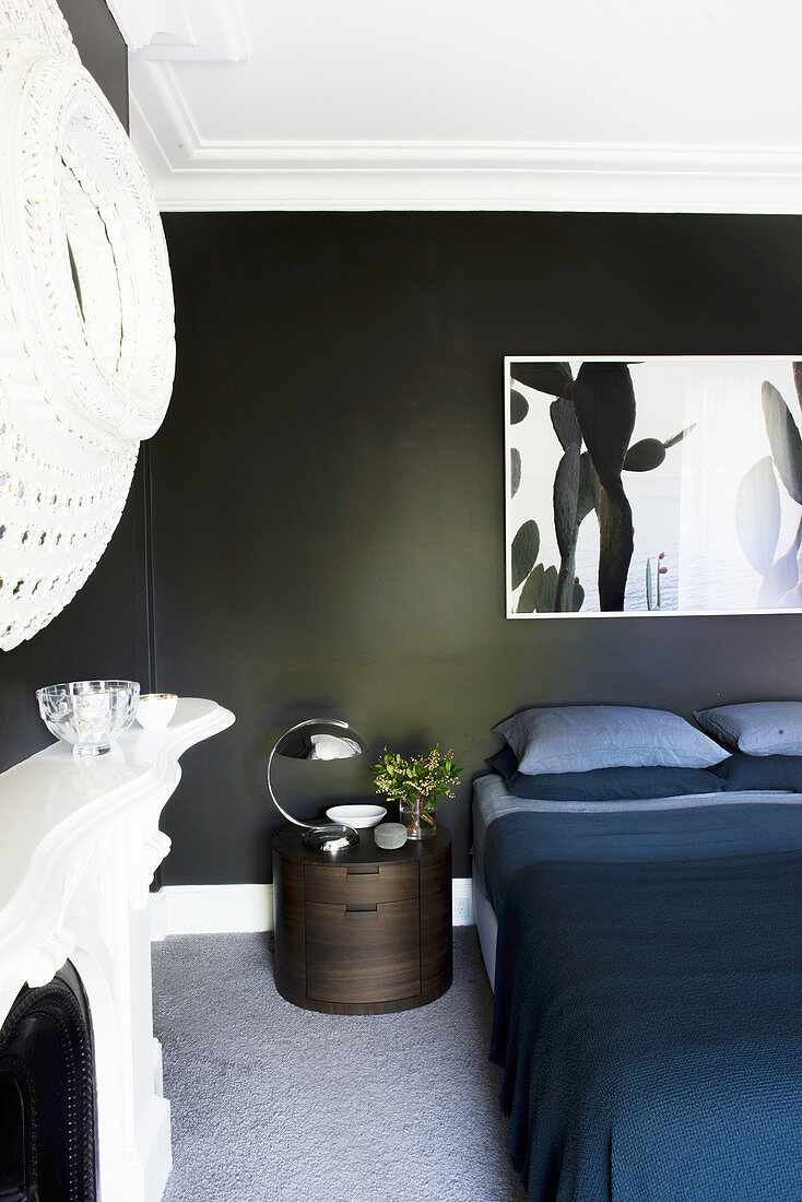 Bild über Doppelbett mit dunkelblauer Tagesdecke, Nachttisch, Kamin und Spiegel im Schlafzimmer mit schwarzen Wänden