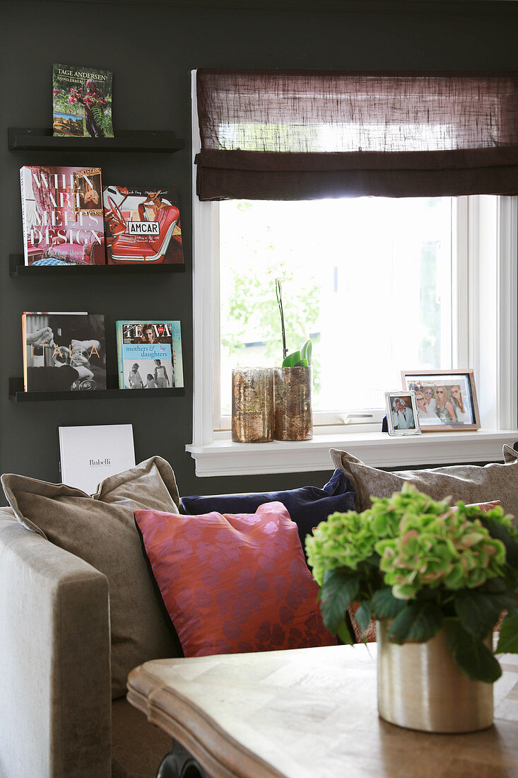 Blick über Couchtisch auf Sofa mit Kissen, Fenster und Bücherregal an dunkler Wand
