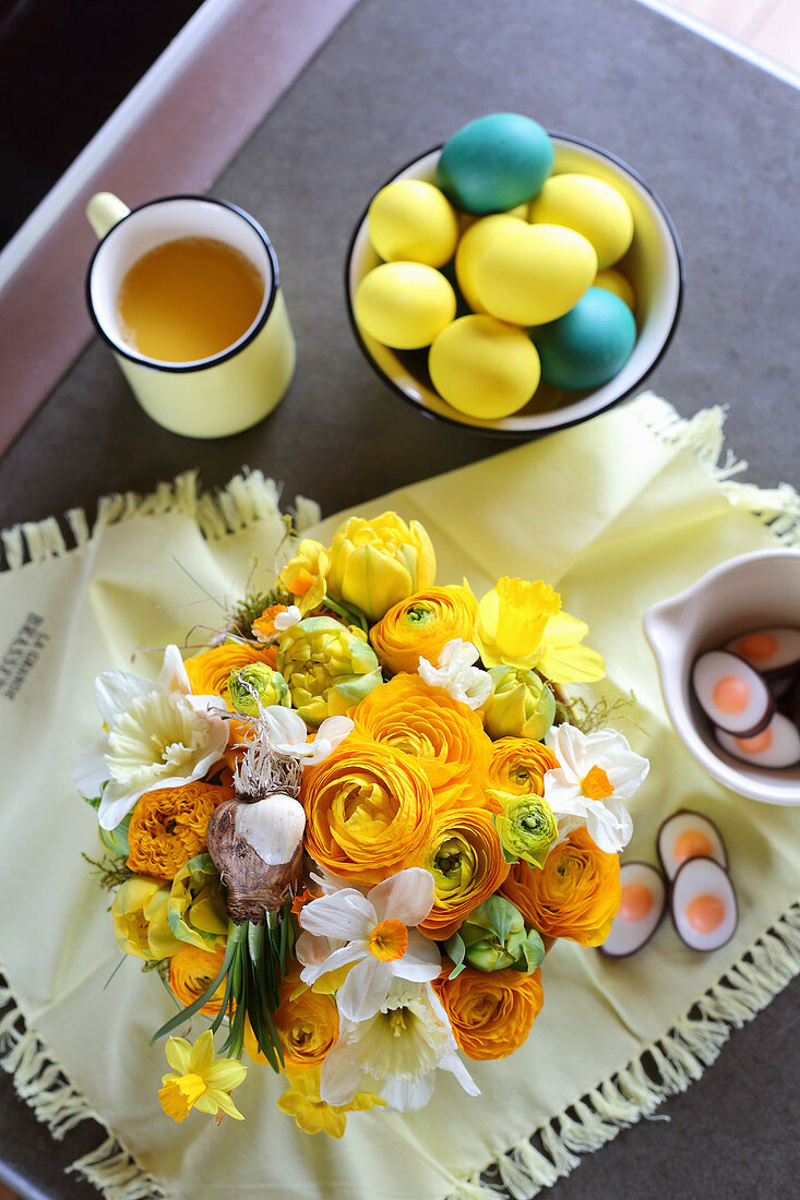 Üppiger Frühlingsstrauß aus Ranunkeln, Tulpen und Tazetten, gelbe und blaue Ostereier und eine Tasse Tee