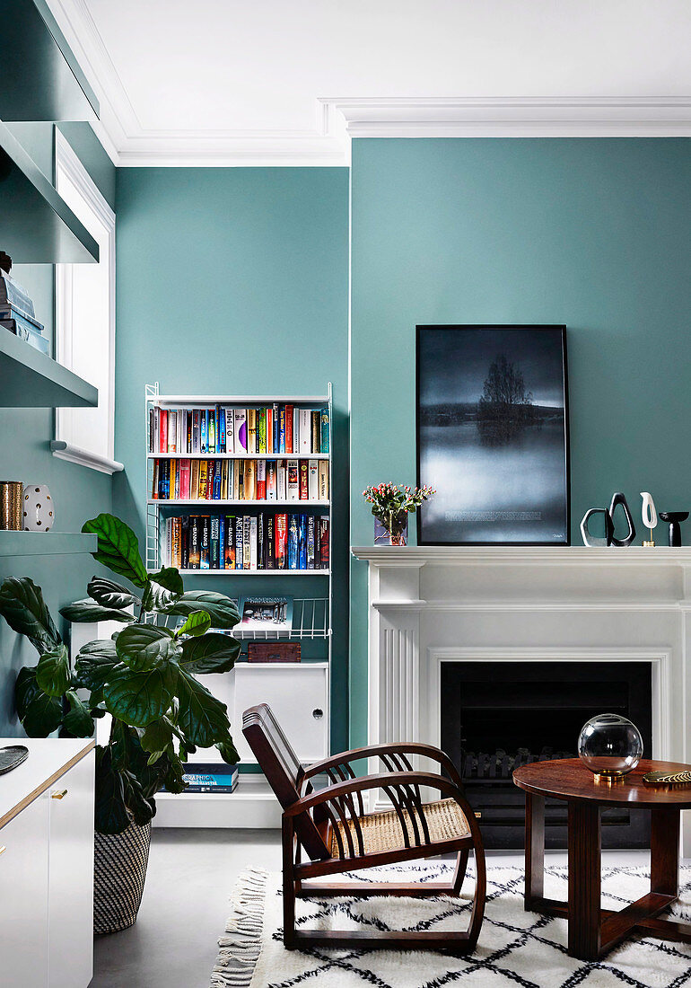 Runder Tisch und Armlehnstuhl vor Kamin, im Hintergrund Bücherregal im Wohnzimmer mit petrolfarbenem Wand