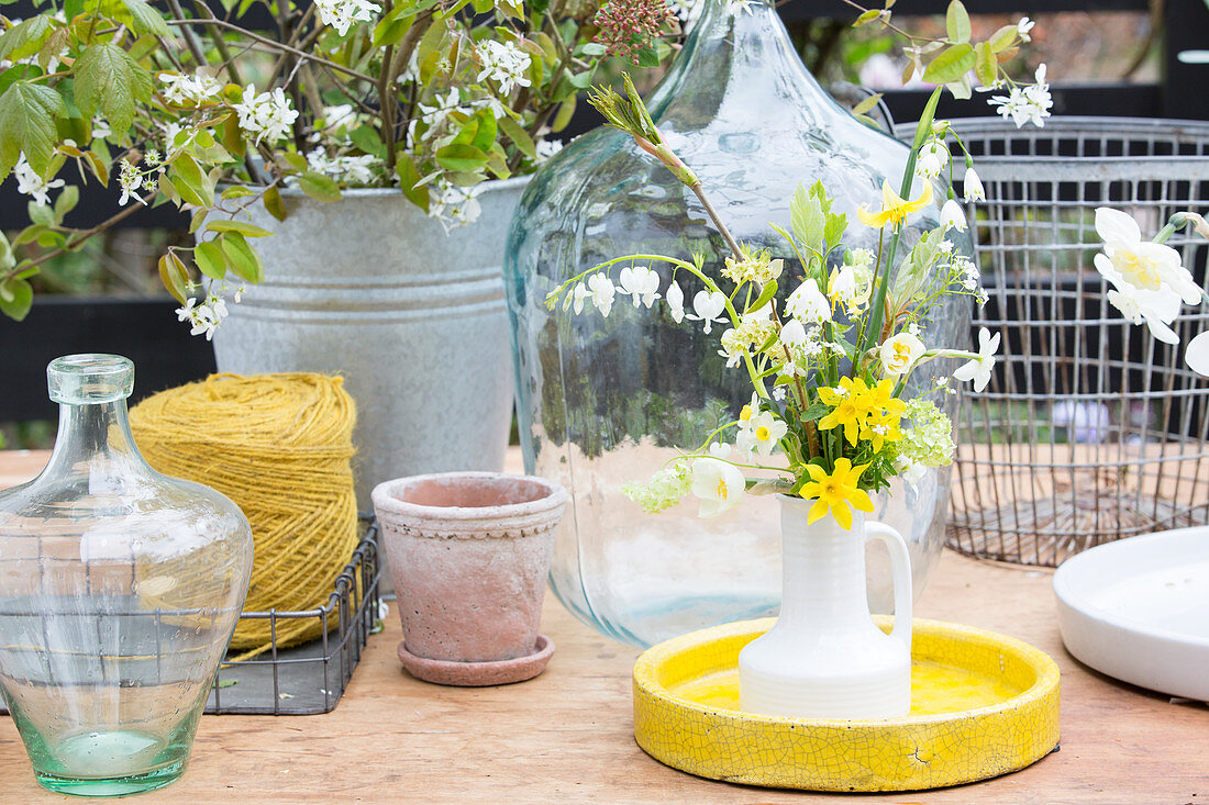 Narzissenstrauß in einer Krugvase, Ballonflaschen, Bindfaden, Pflanzentopf und Zinkeimer auf Gartentisch