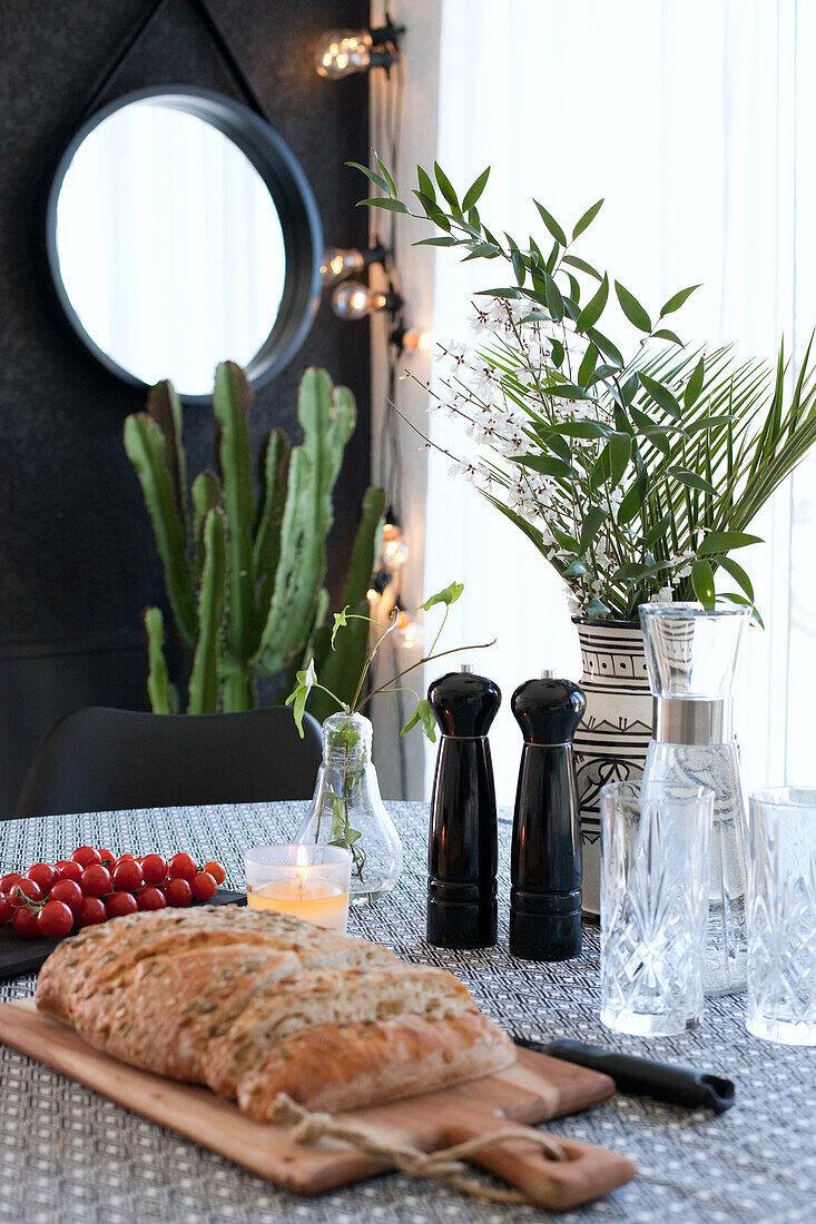 Loaf on cutting board, glasses, salt and pepper mills and vase of leafy branches on kitchen table