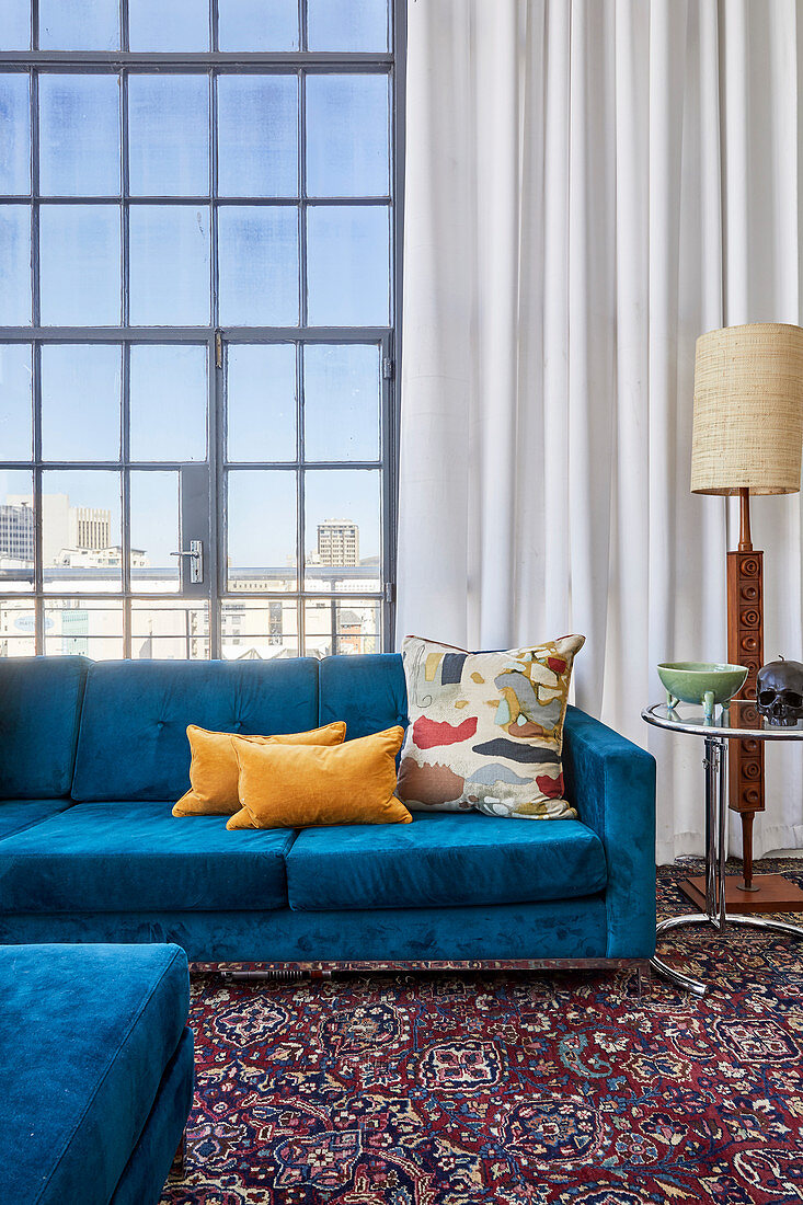 Scatter cushions on bright blue sofa, side table and standard lamp against glass wall in high-ceilinged interior
