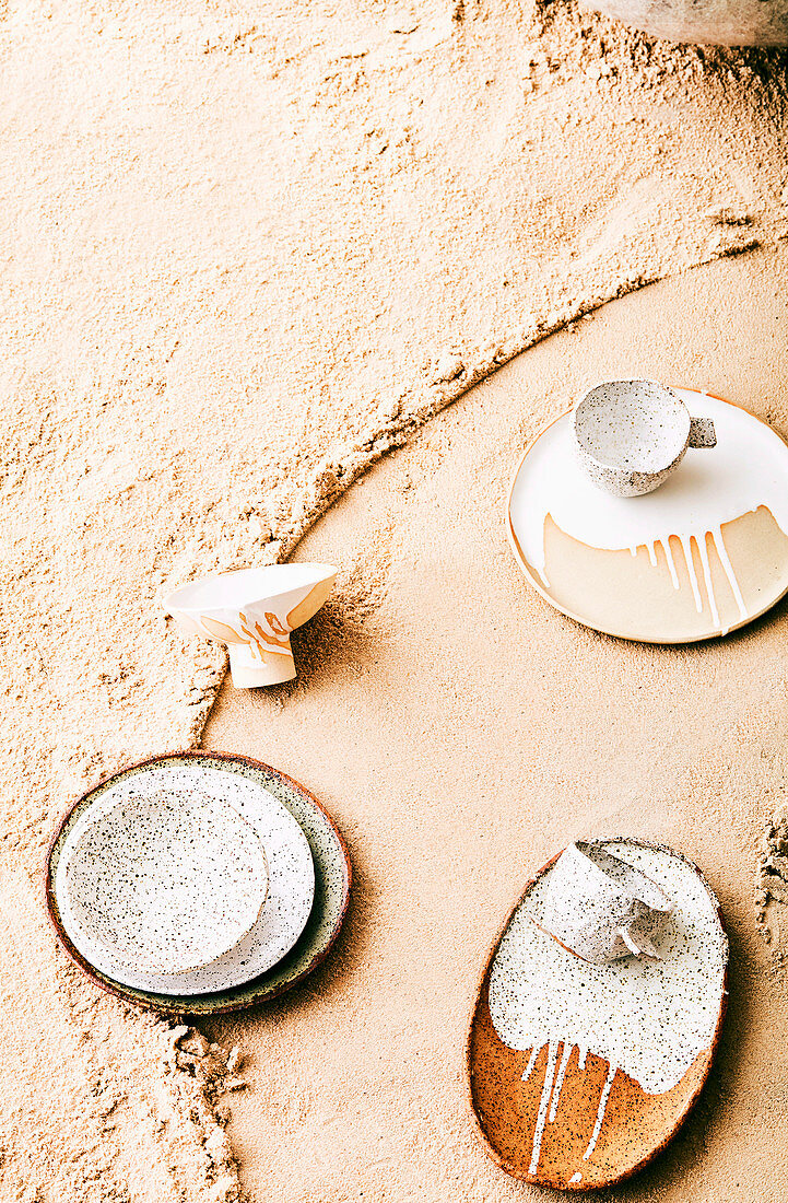 Teller und Tassen mit gesprenkelter und verlaufener Glasur auf Sand