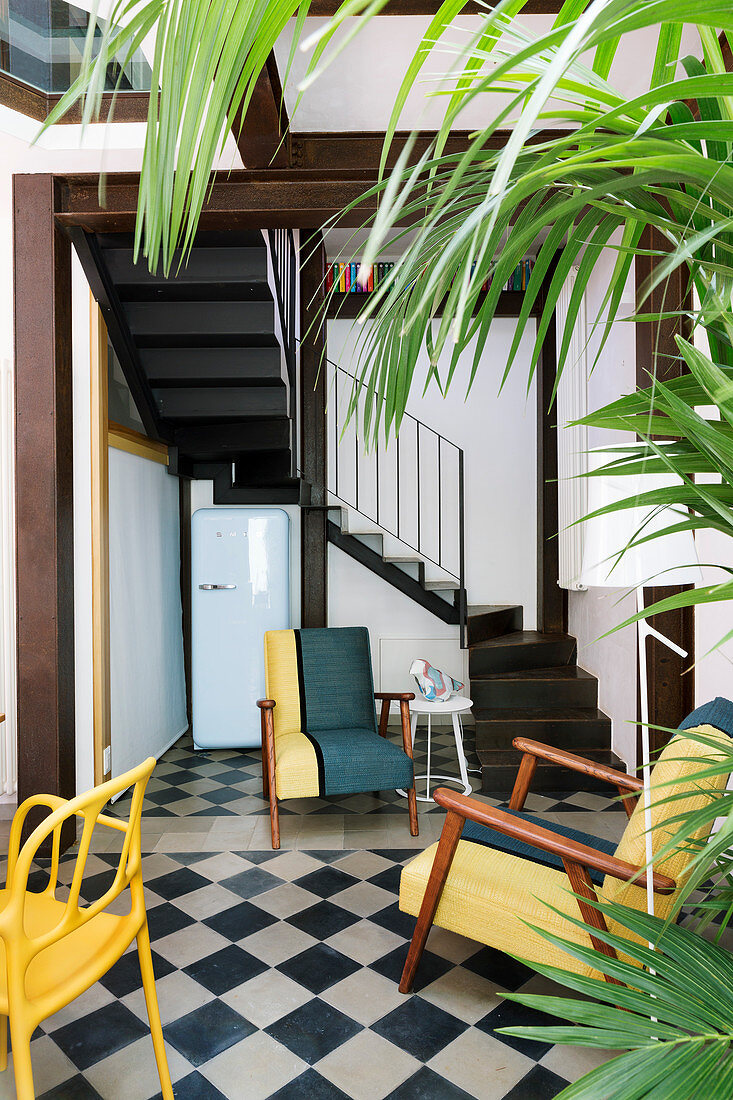 Colourful seating on chequered floor and houseplants in interior with staircase in background