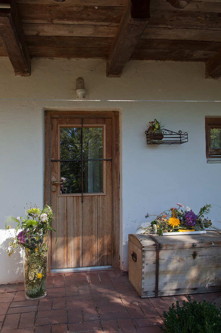 Glasvase mit Herbstblumen und Blumenpotpourri auf Holztruhe am Hauseingang