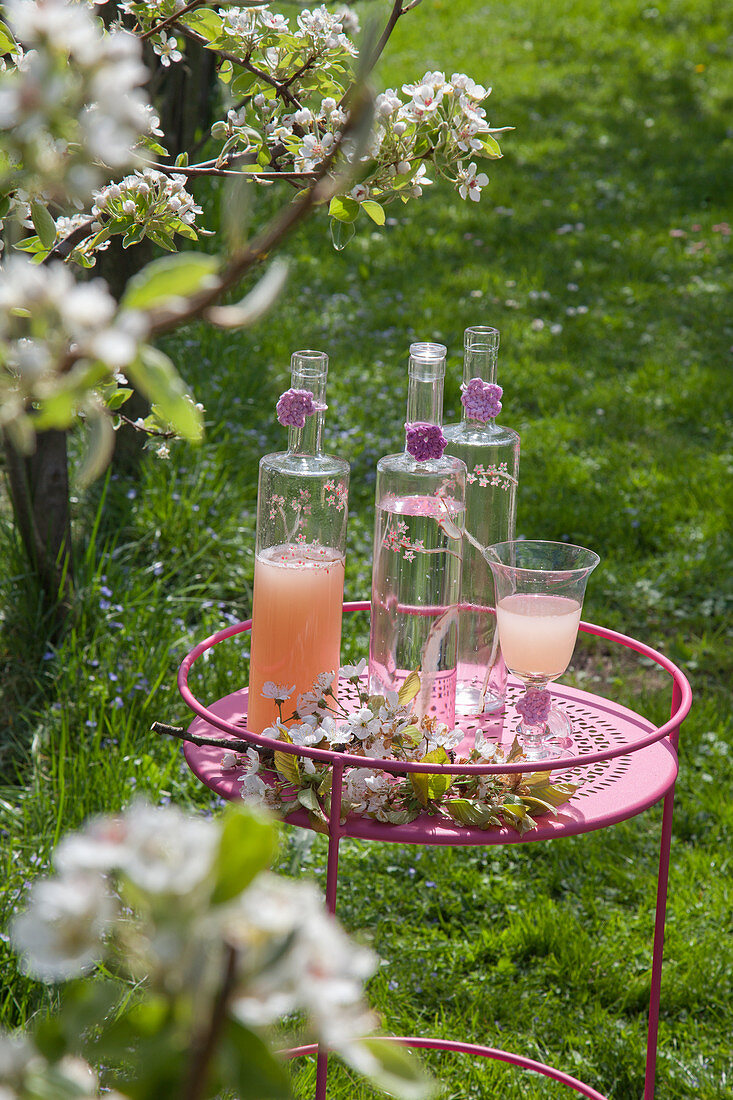 Erfrischungsgetränke in Flaschen und zarte Kirschblüten auf rundem Beistelltisch im Garten