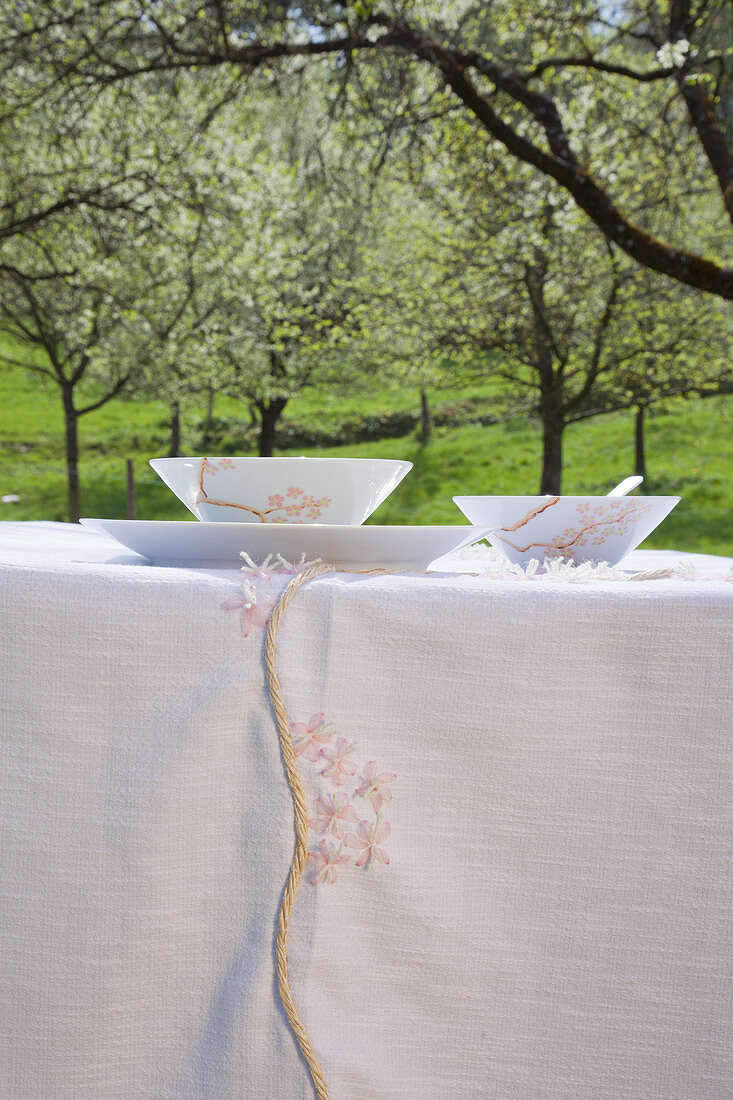 Crockery and handmade tablecloth with cherry blossom patterns on table below flowering cherry tree in garden