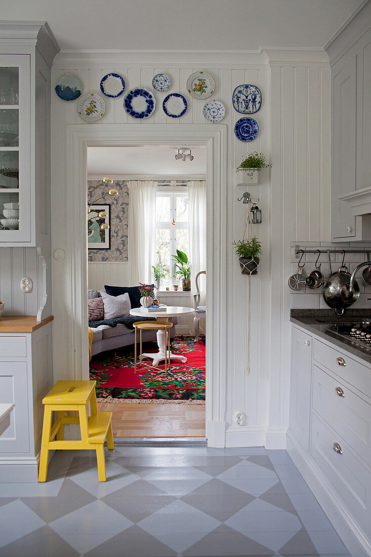 Gelber Tritthocker in Einbauküche mit bemaltem Dielenboden, Wandteller an Holzverkleidung über Türrahmen, Blick in Wohnzimmer