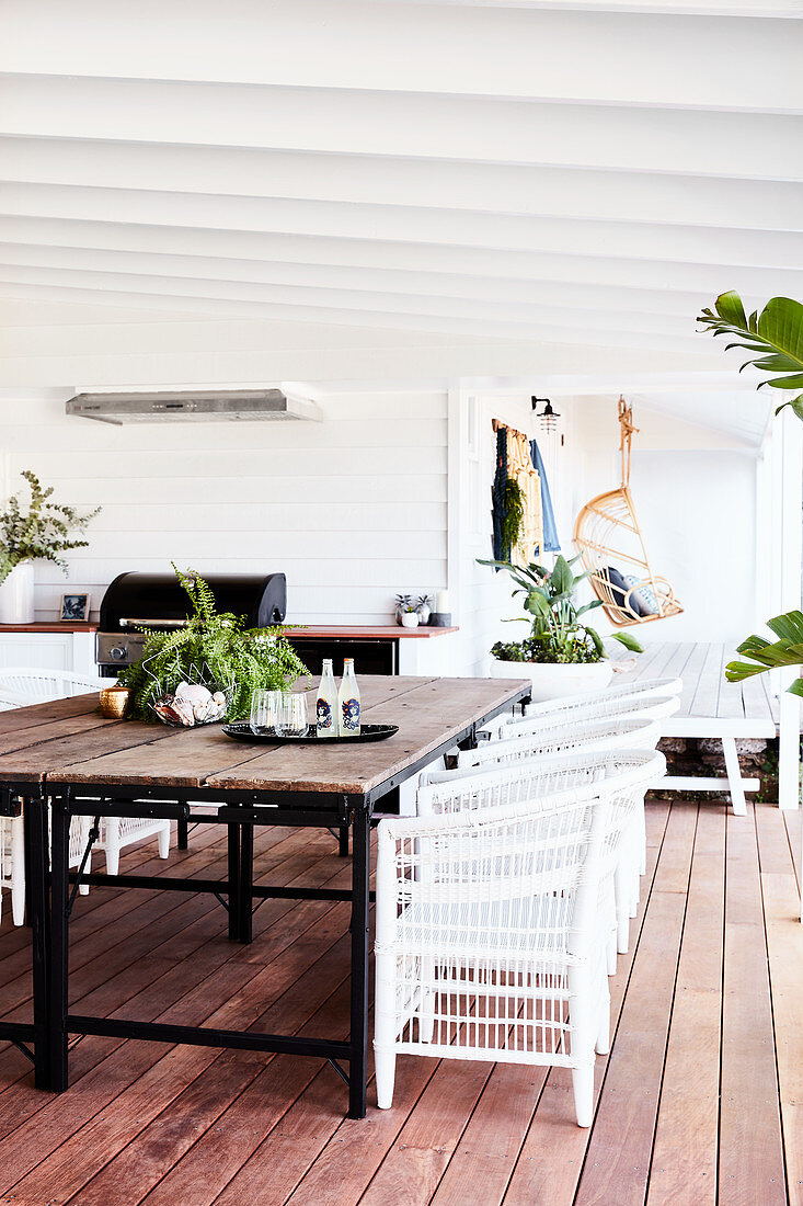 Esstisch mit rustikaler Holzplatte und weißen Rattanstühlen auf überdachter Terrasse