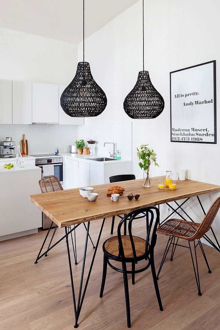 Dining table and various chairs below black pendant lamps in open-plan kitchen