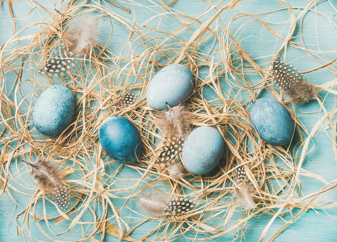 Blue-painted Easter eggs in hay and feathers (top view)