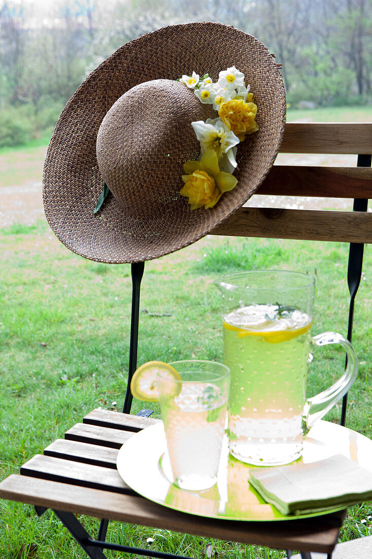 Zitronenlimonade auf Gartenstuhl und Sommerhut mit Blütendeko